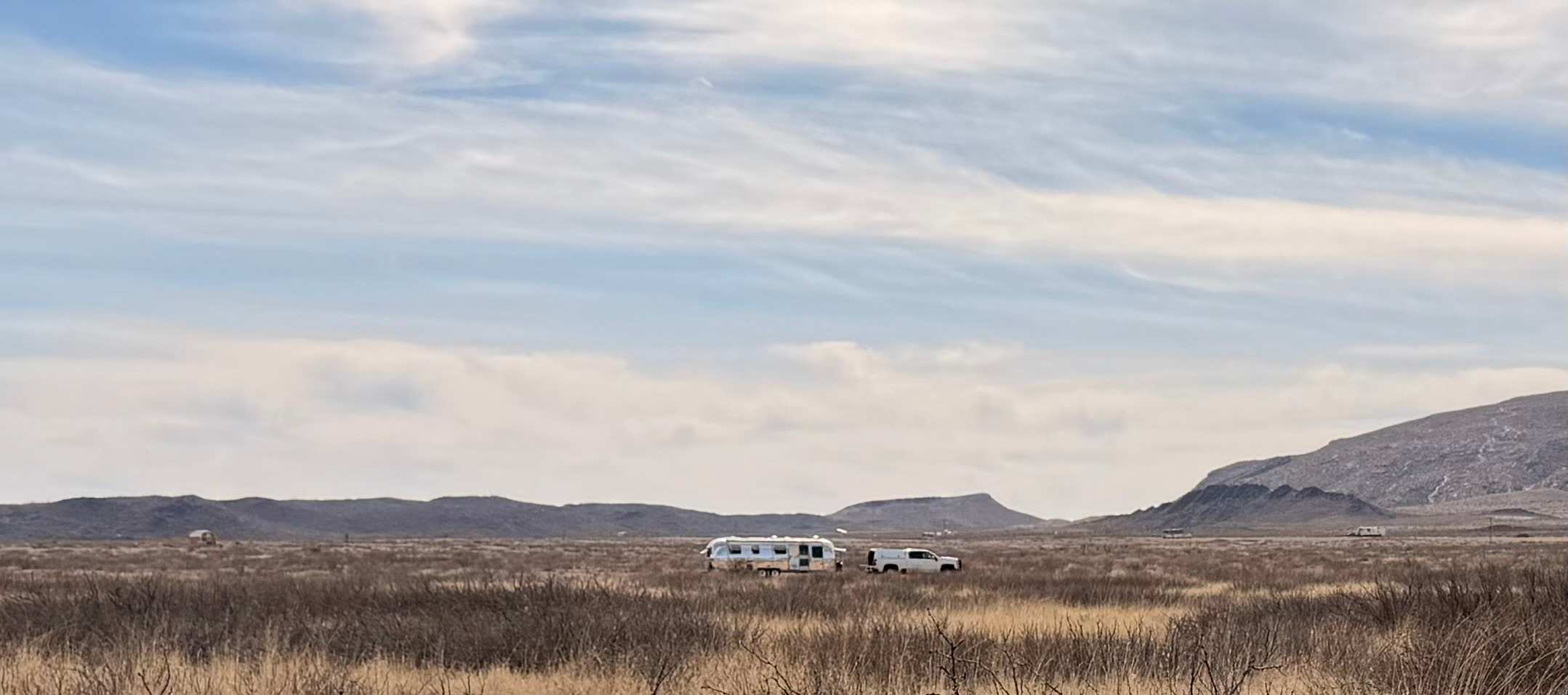 Wide panorama of golden desert grassland with an Airstream and pickup parked beneath distant mountains and a vast pale sky.