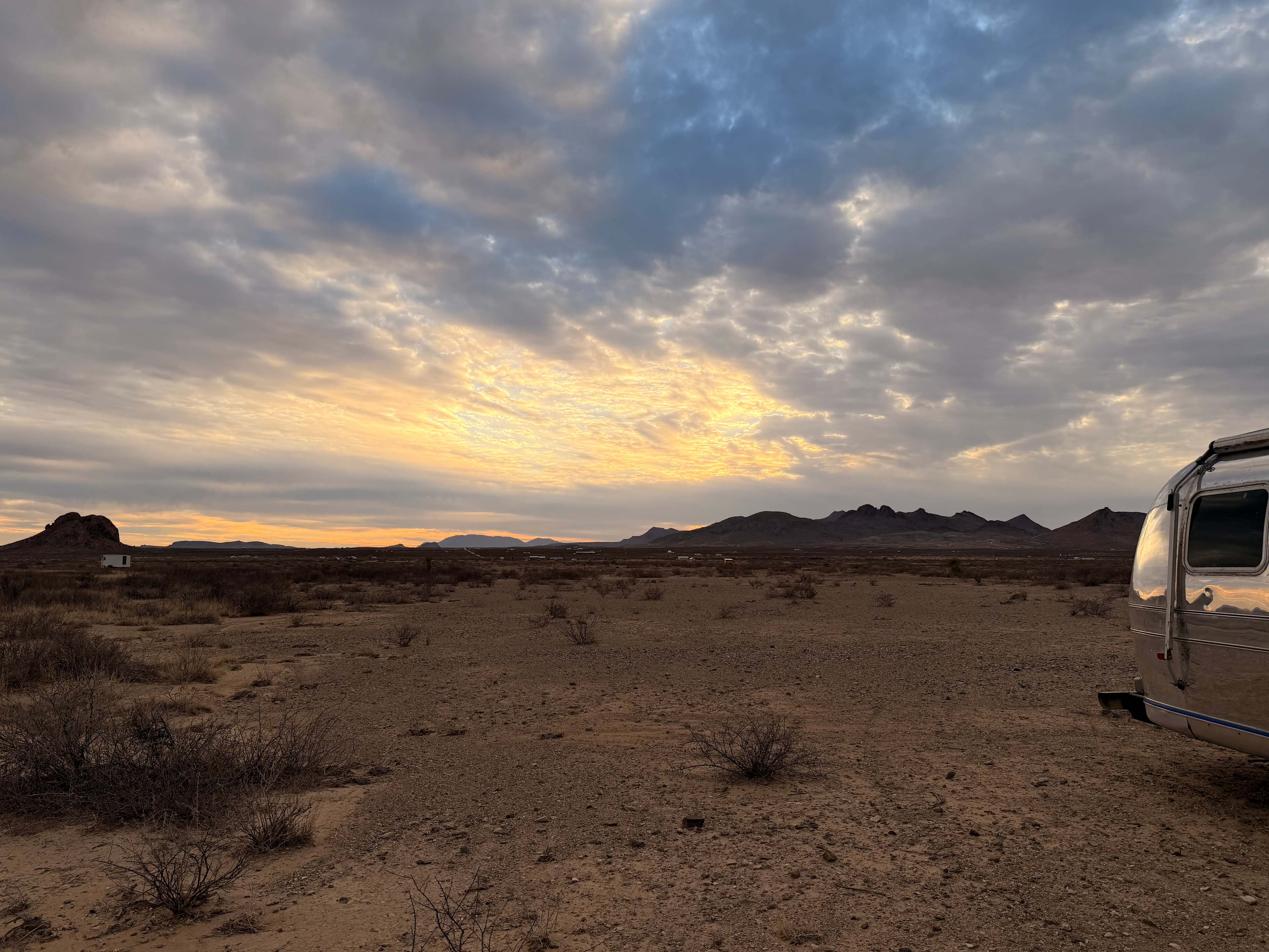 Dawn breaking gold through heavy cloud over the desert, an Airstream parked at the far right of frame.