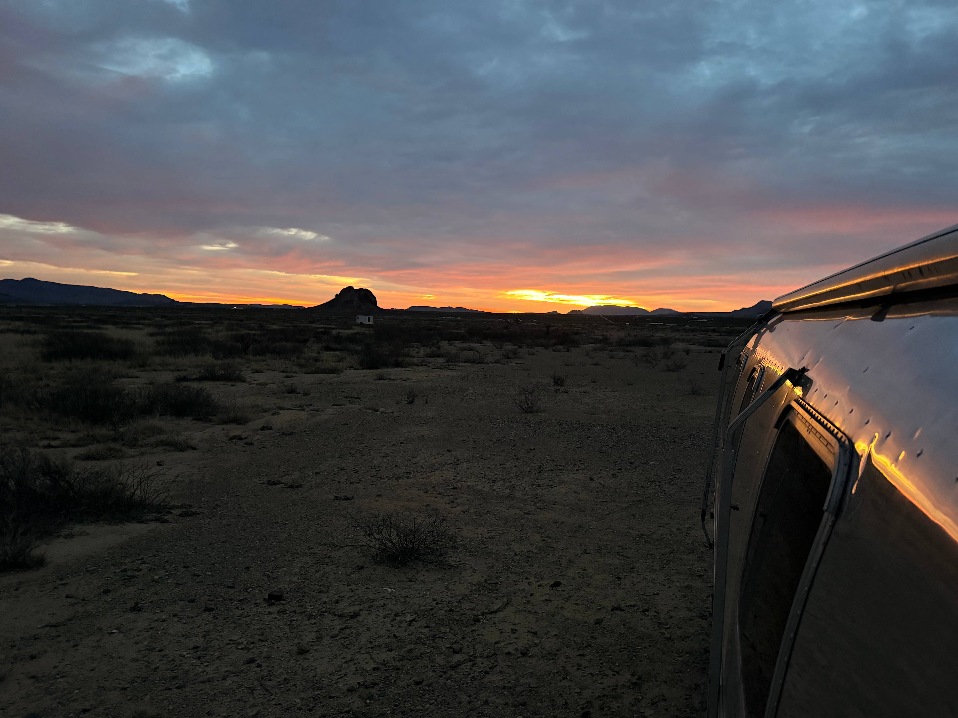Deep orange and violet sunset over the desert, the curve of a silver Airstream catching the last light.